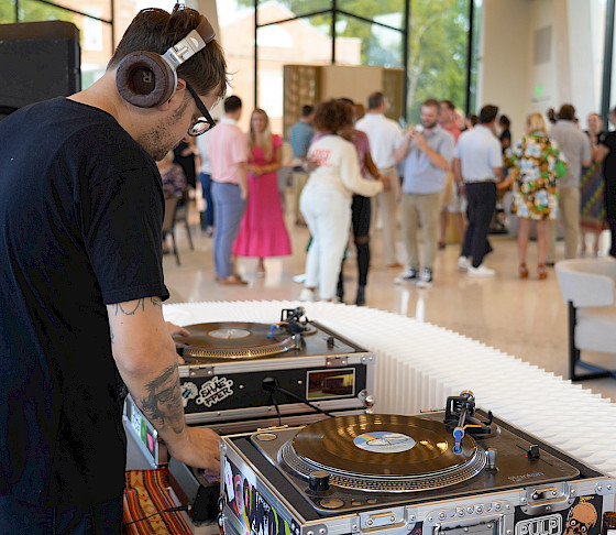 Photo of a DJ mixing records on a set of turntables with groups of people mingling and dancing at a party in the background.