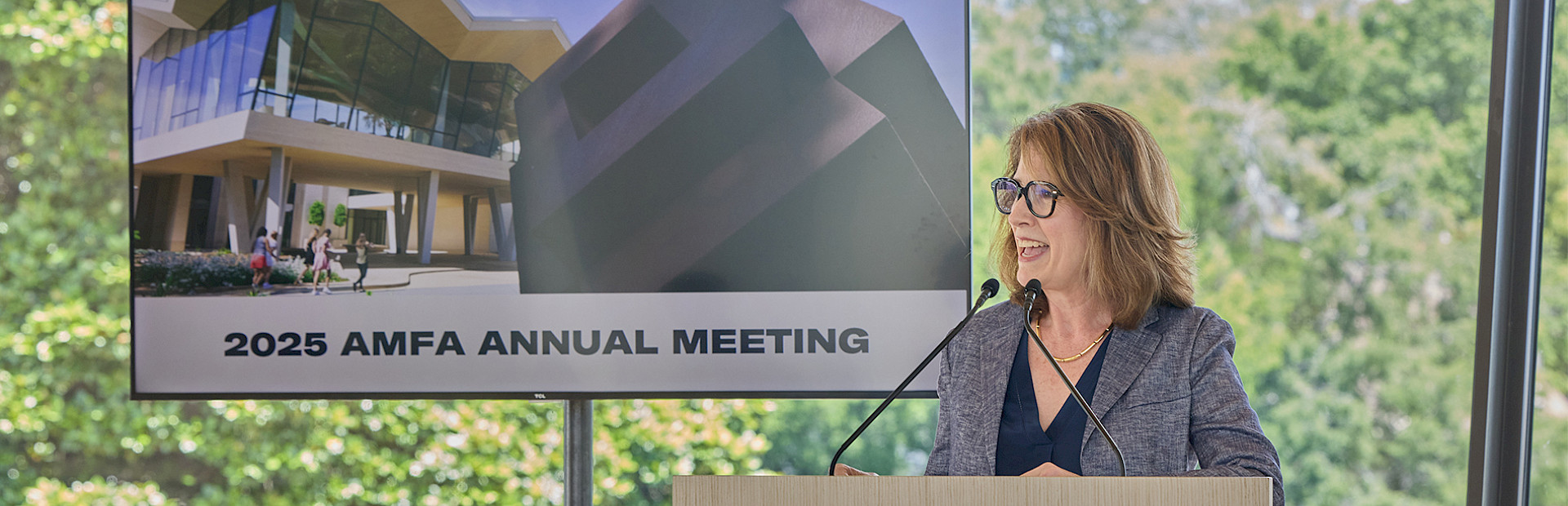 Photo of Dr. Victoria Ramirez speaking at a podium with a screen behind her displaying a photo of the Arkansas Museum of Fine Arts with the words 2025 AMFA Annual Meeting.