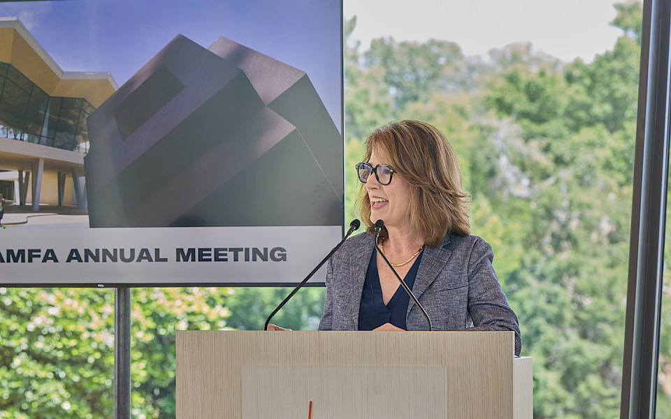 Photo of Dr. Victoria Ramirez speaking at a podium with a screen behind her displaying a photo of the Arkansas Museum of Fine Arts with the words 2025 AMFA Annual Meeting.