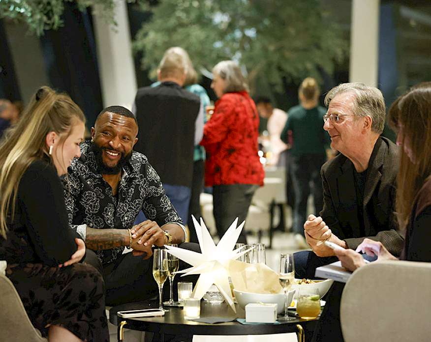Photo of two men and two women sitting an a table drinking champagne with other people in the background.